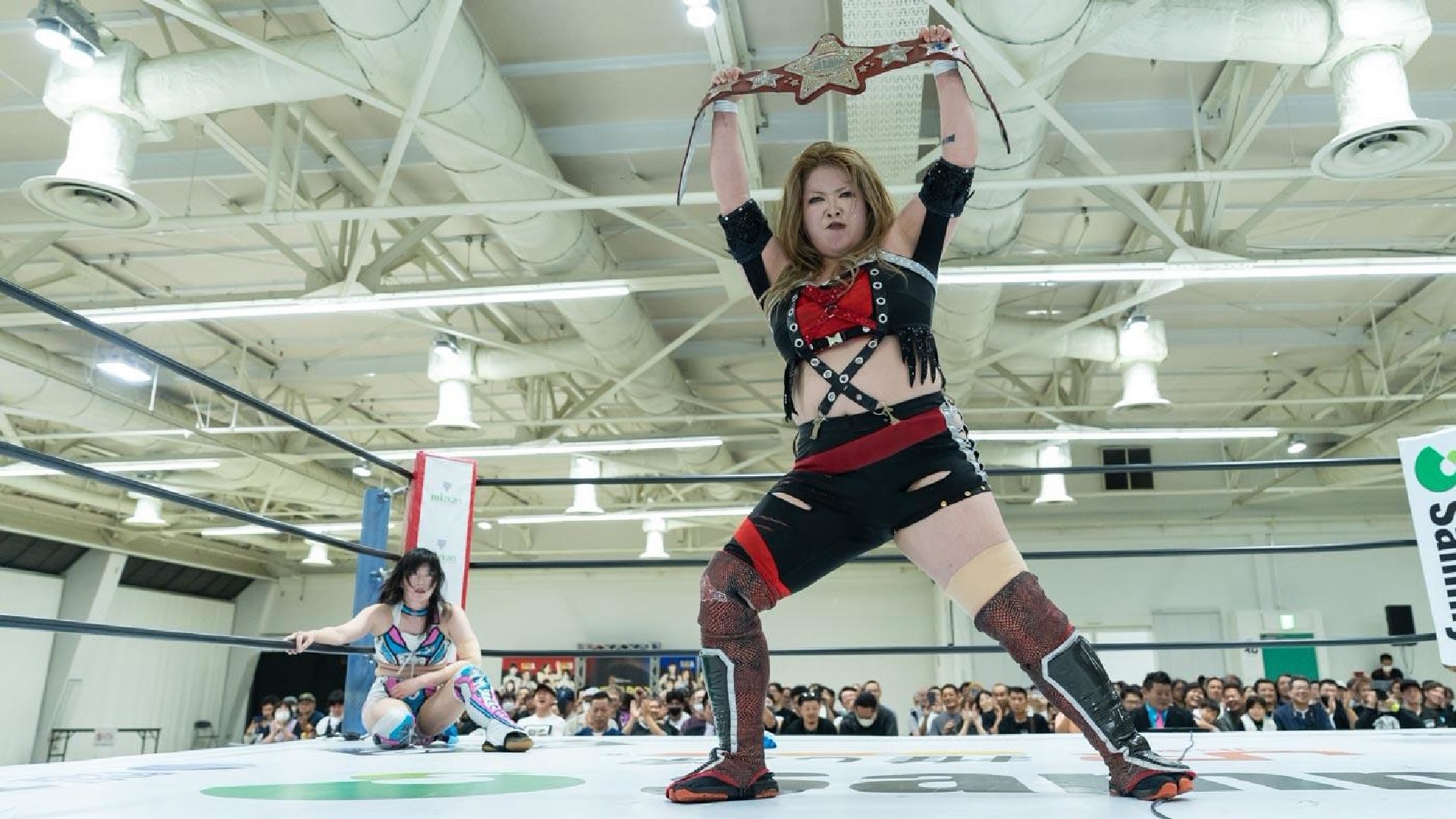 Wrestler in black-and-red gear raises a championship belt overhead inside a wrestling ring, crowd watching from the stands.