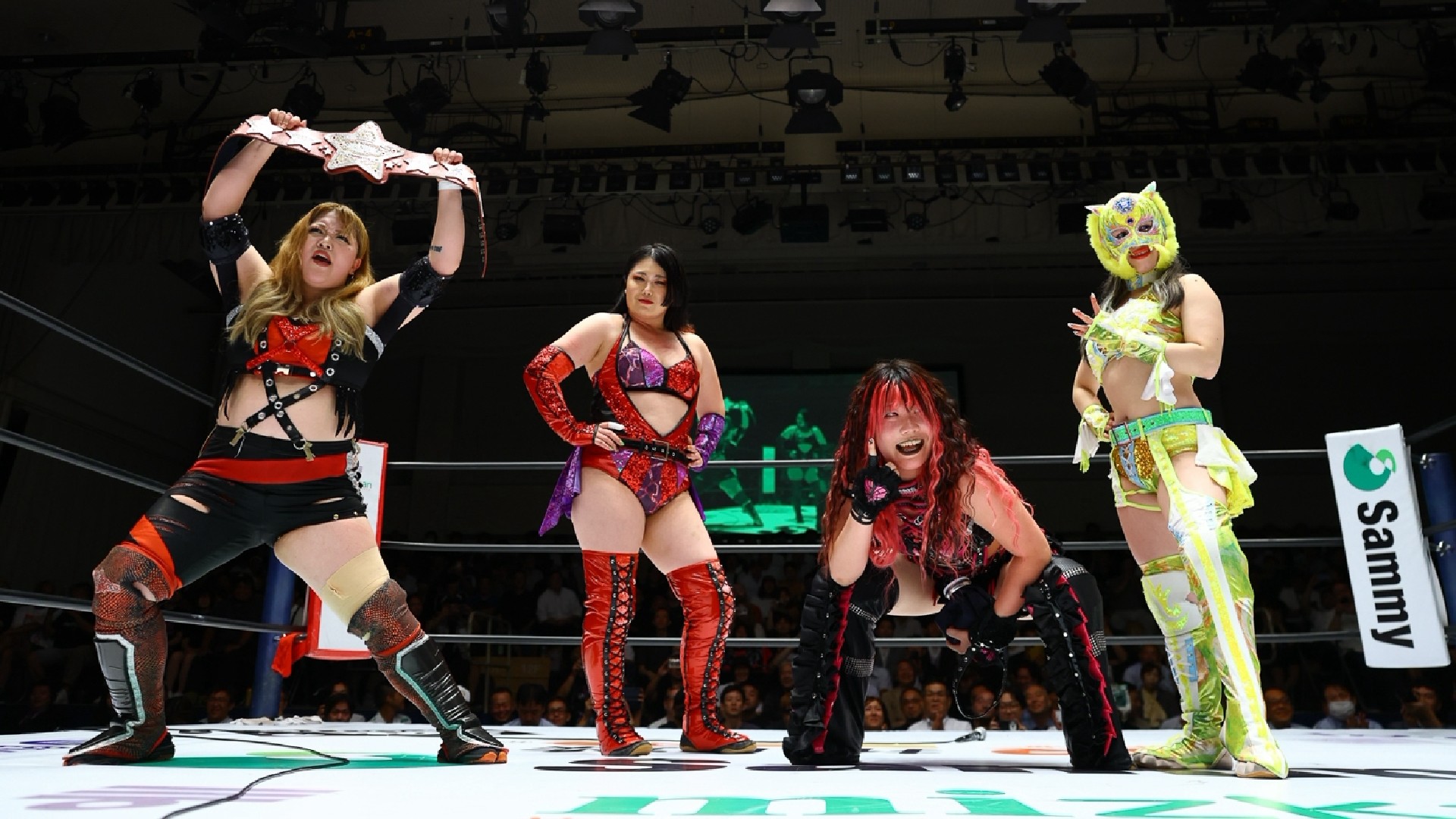 Four female wrestlers pose in a ring, colorful outfits, as the crowd watches from the stands.