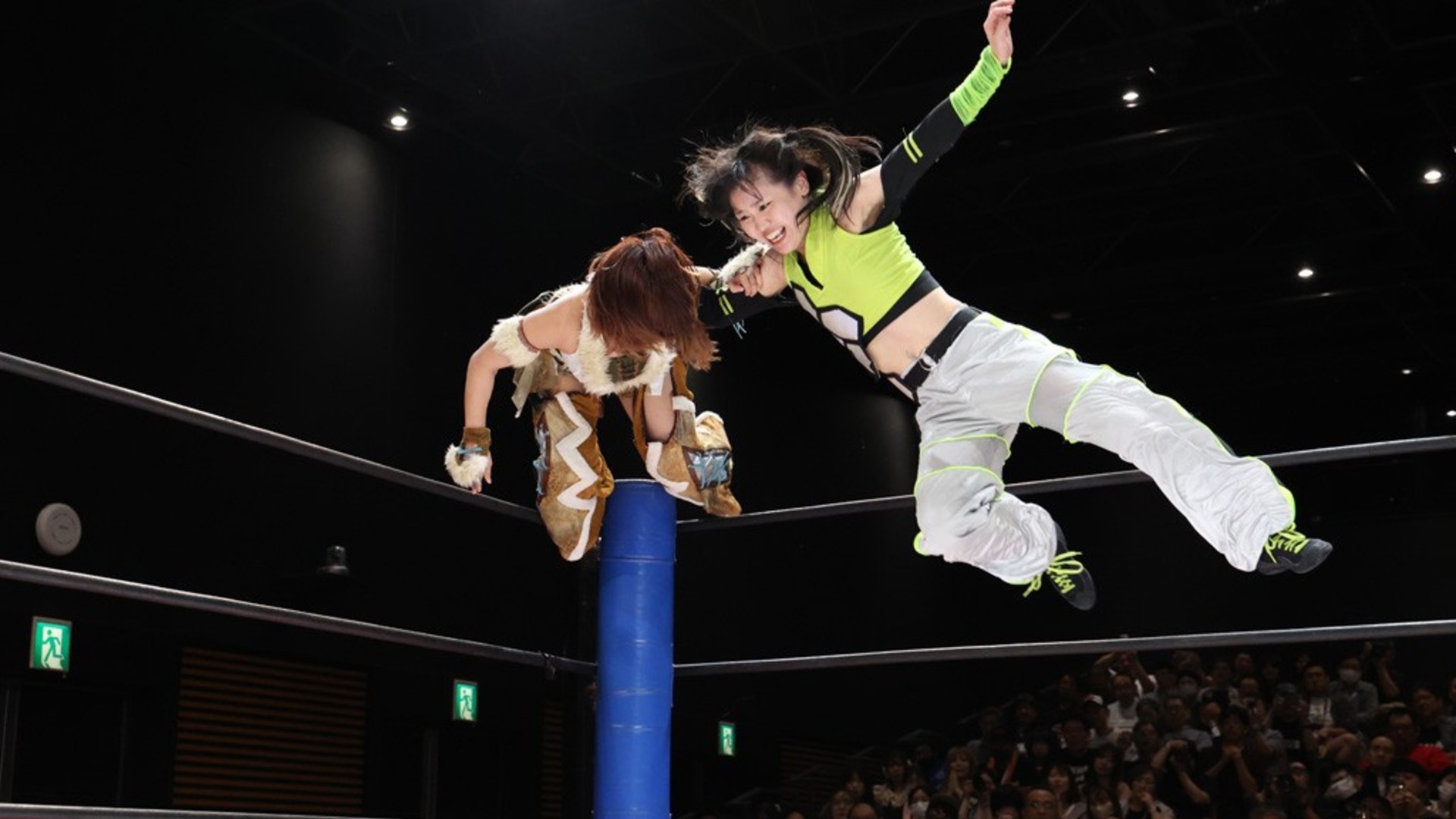 Wrestlers in a ring: one athlete in neon green and gray gear soars mid-air from the ropes toward a partner on the turnbuckle, crowd watching.