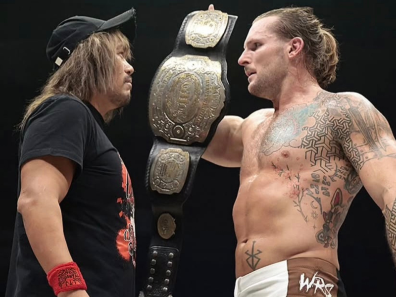 Two wrestlers face off with a large championship belt between them, preparing for a match.