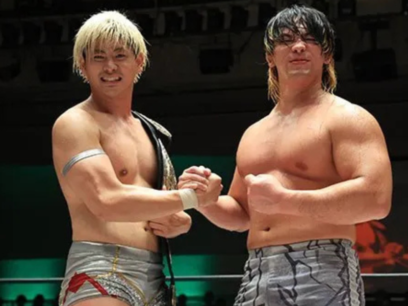 Two shirtless wrestlers shake hands in a ring after a match, one wearing a championship belt over his shoulder and silver trunks with red details.