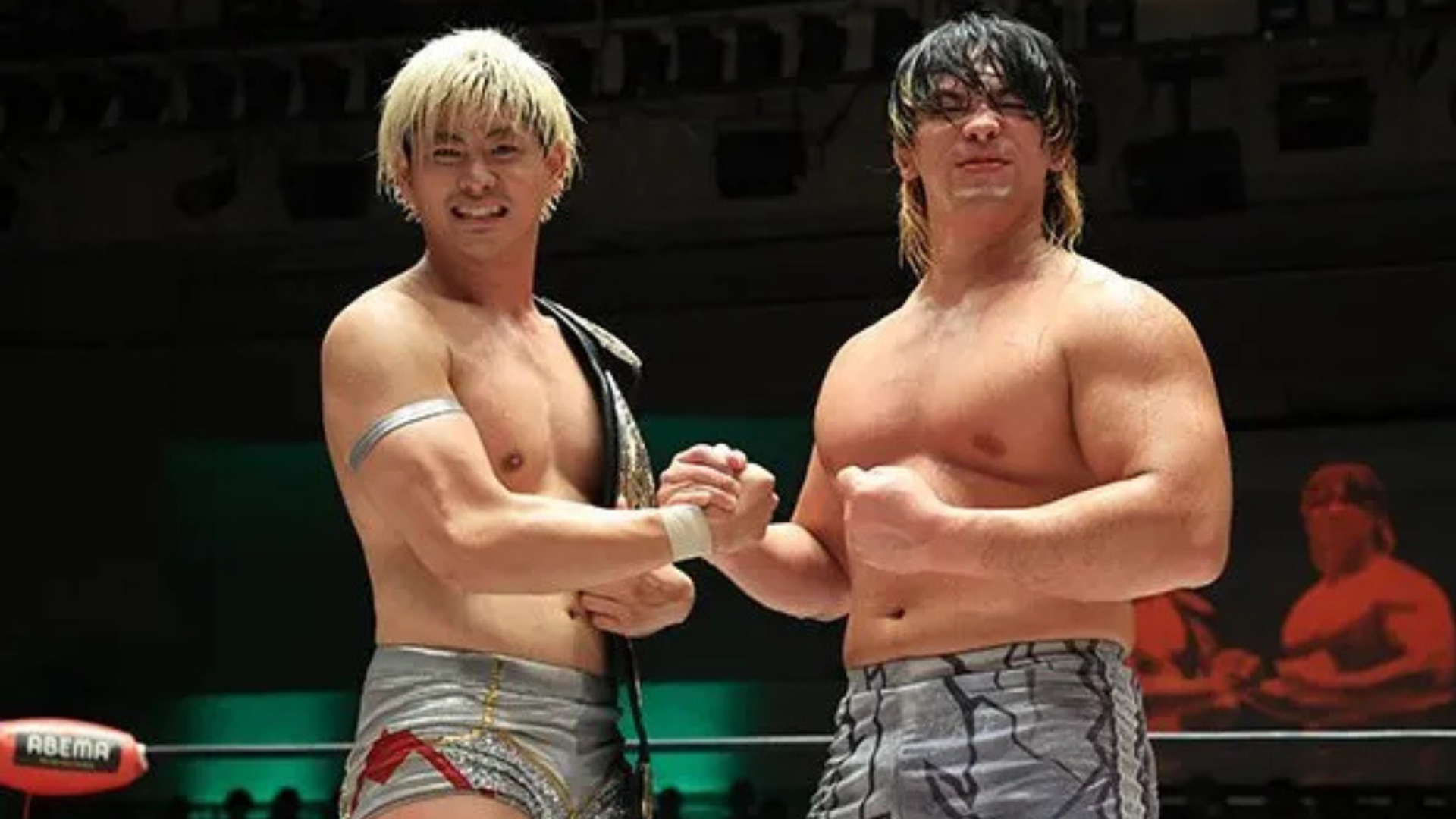 Two shirtless wrestlers shake hands in a ring after a match, one wearing a championship belt over his shoulder and silver trunks with red details.