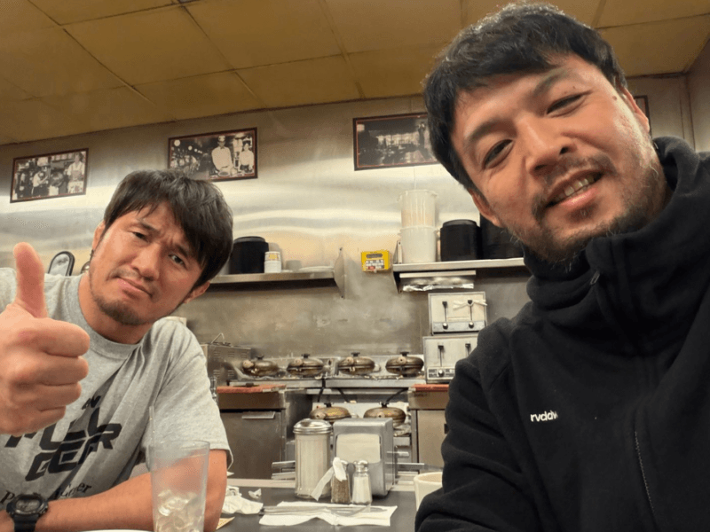 Two men in a kitchen take a selfie, one giving a thumbs-up to the camera.