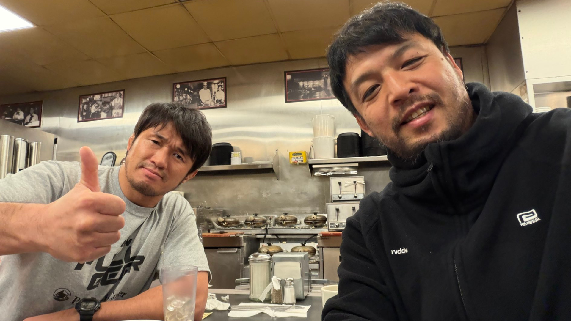 Two men in a kitchen take a selfie, one giving a thumbs-up to the camera.
