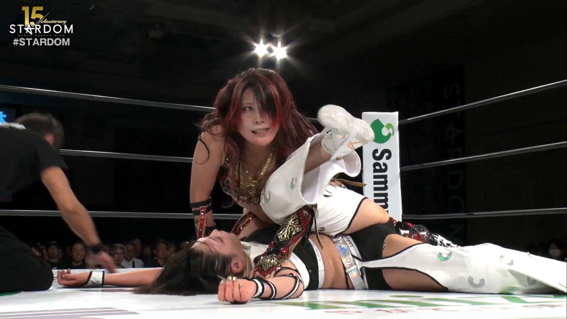 Two female wrestlers grapple on the mat inside a ring; one pins the other as a referee looks on, with Stardom branding in the corner.