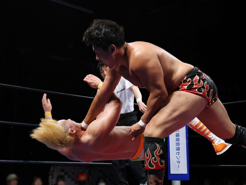 Two muscular wrestlers clash in the ring during a match, one delivering a throw to the other who is mid-air.