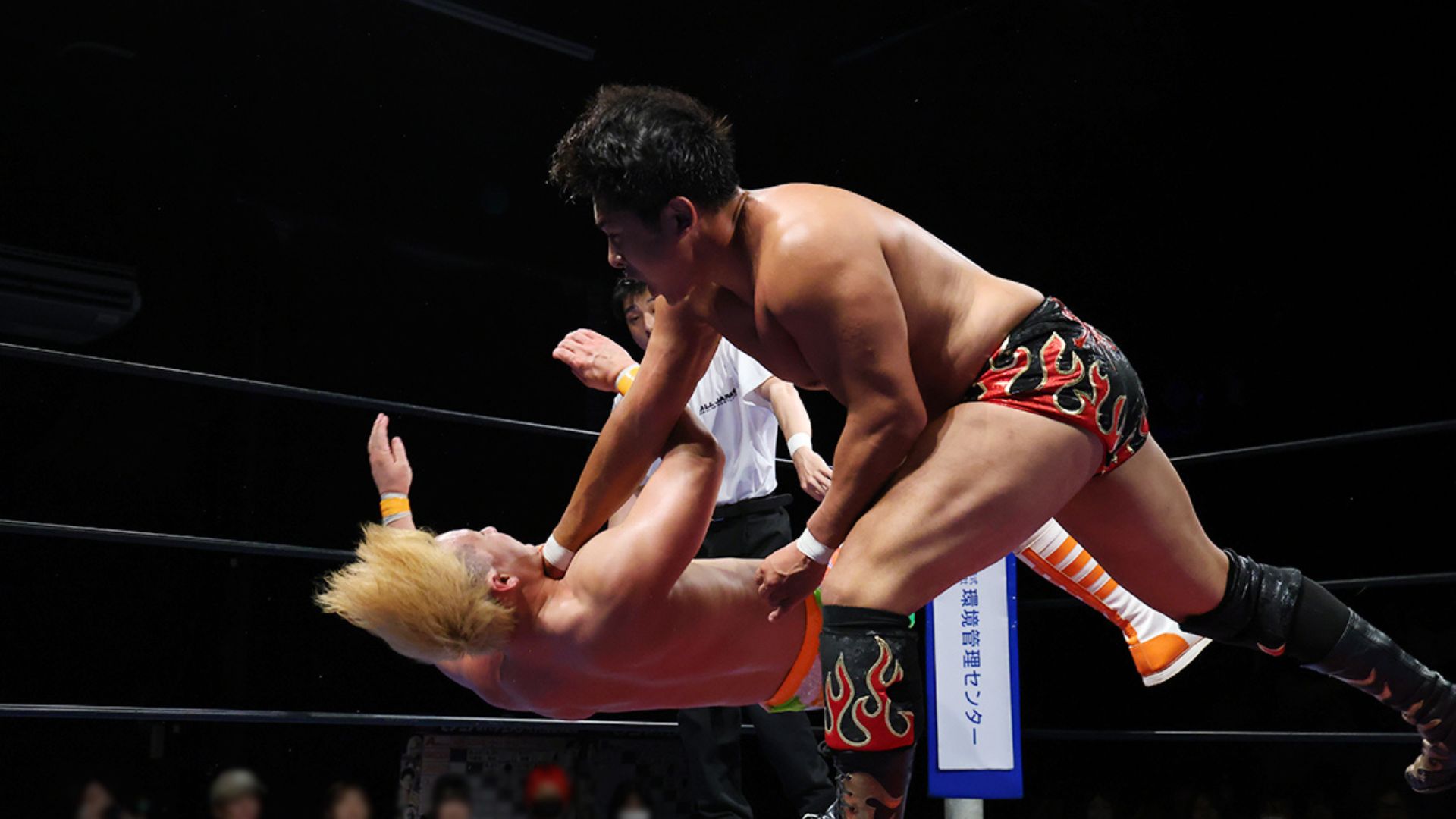 Two muscular wrestlers clash in the ring during a match, one delivering a throw to the other who is mid-air.