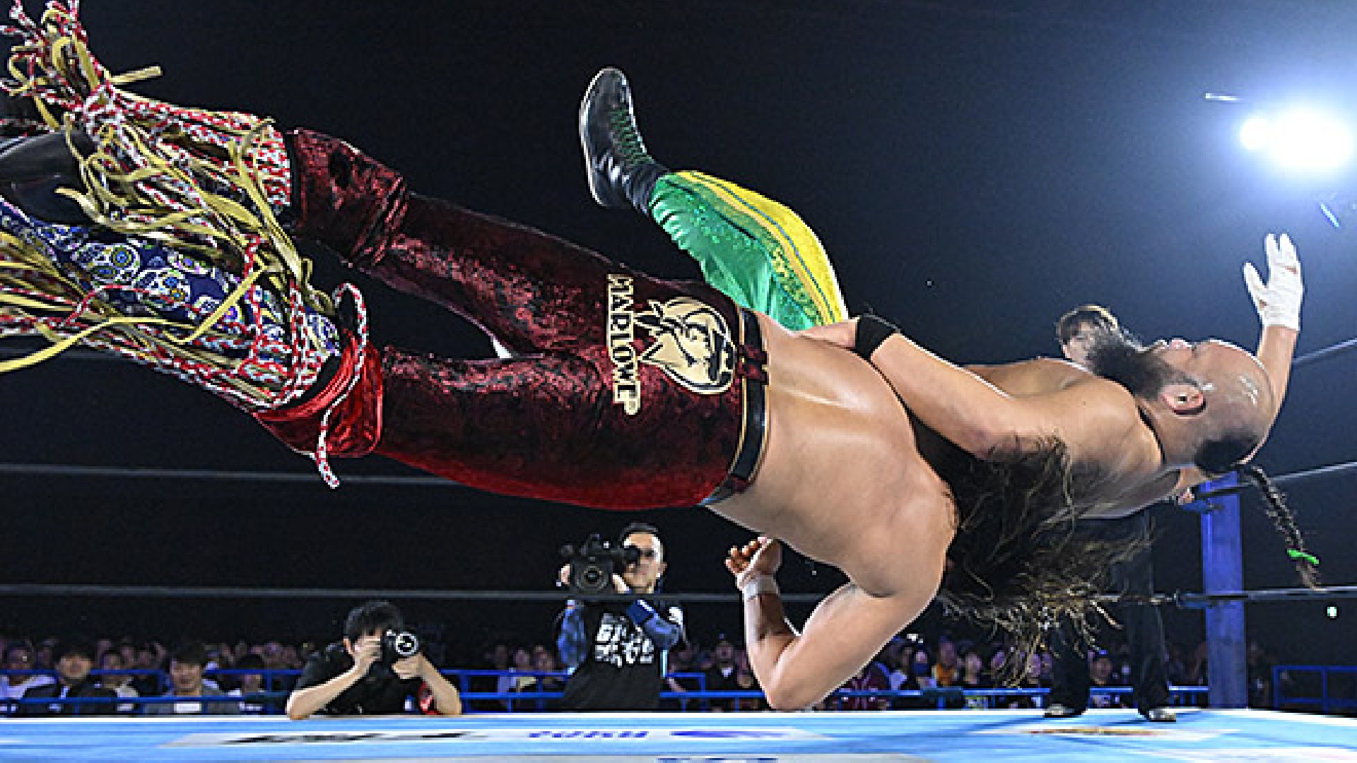 High‑flying wrestling move: two competitors collide mid‑air in the ring as photographers and a cheering crowd look on.