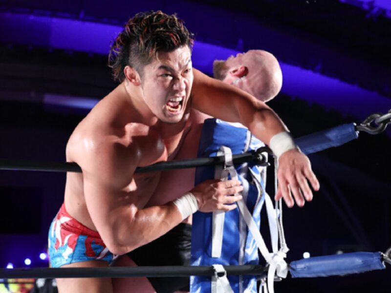 Wrestler in red shorts applies a headlock on an opponent leaning on the ring ropes during a match in a dark arena.