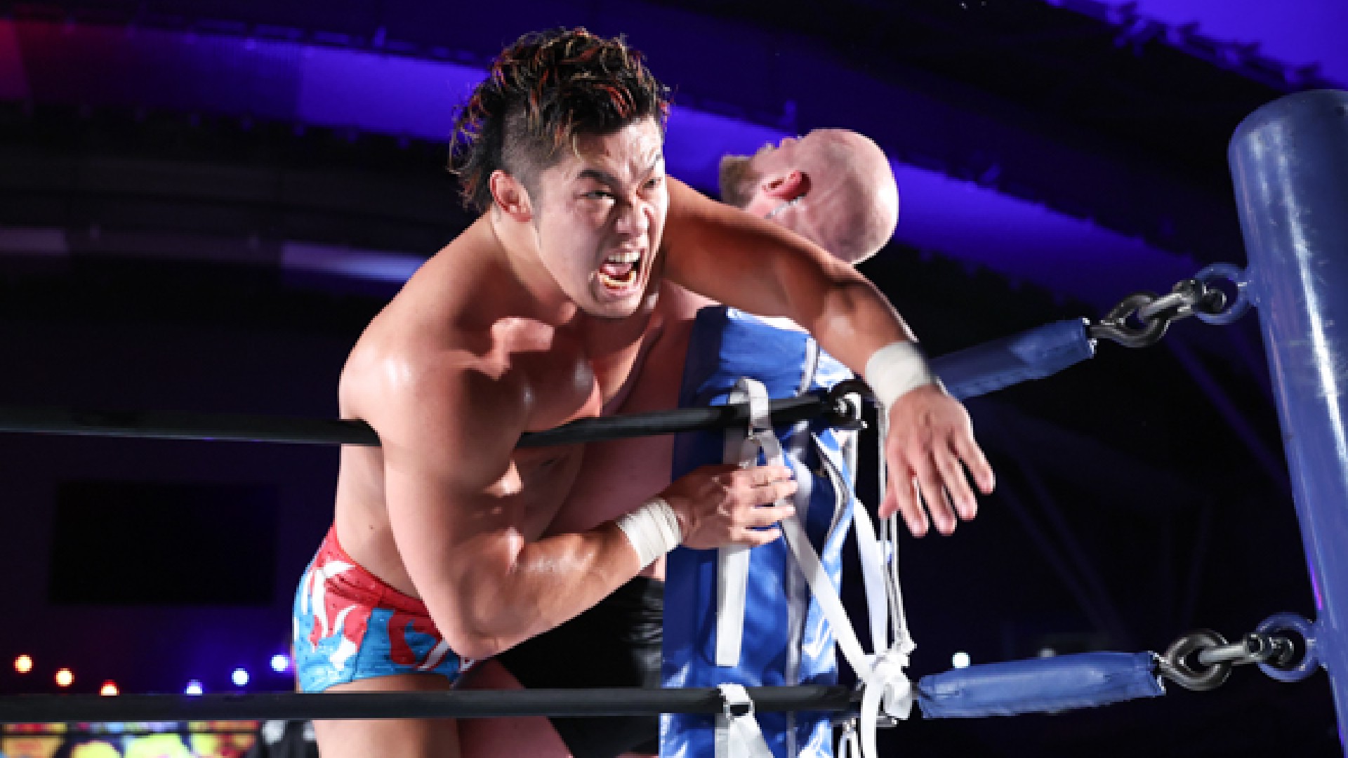 Wrestler in red shorts applies a headlock on an opponent leaning on the ring ropes during a match in a dark arena.