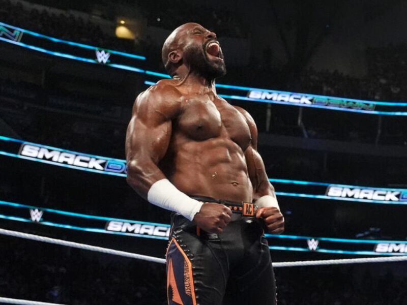 Muscular male wrestler in a WWE ring, fists clenched and shouting, wearing black/orange trunks and white wristbands, celebration pose.