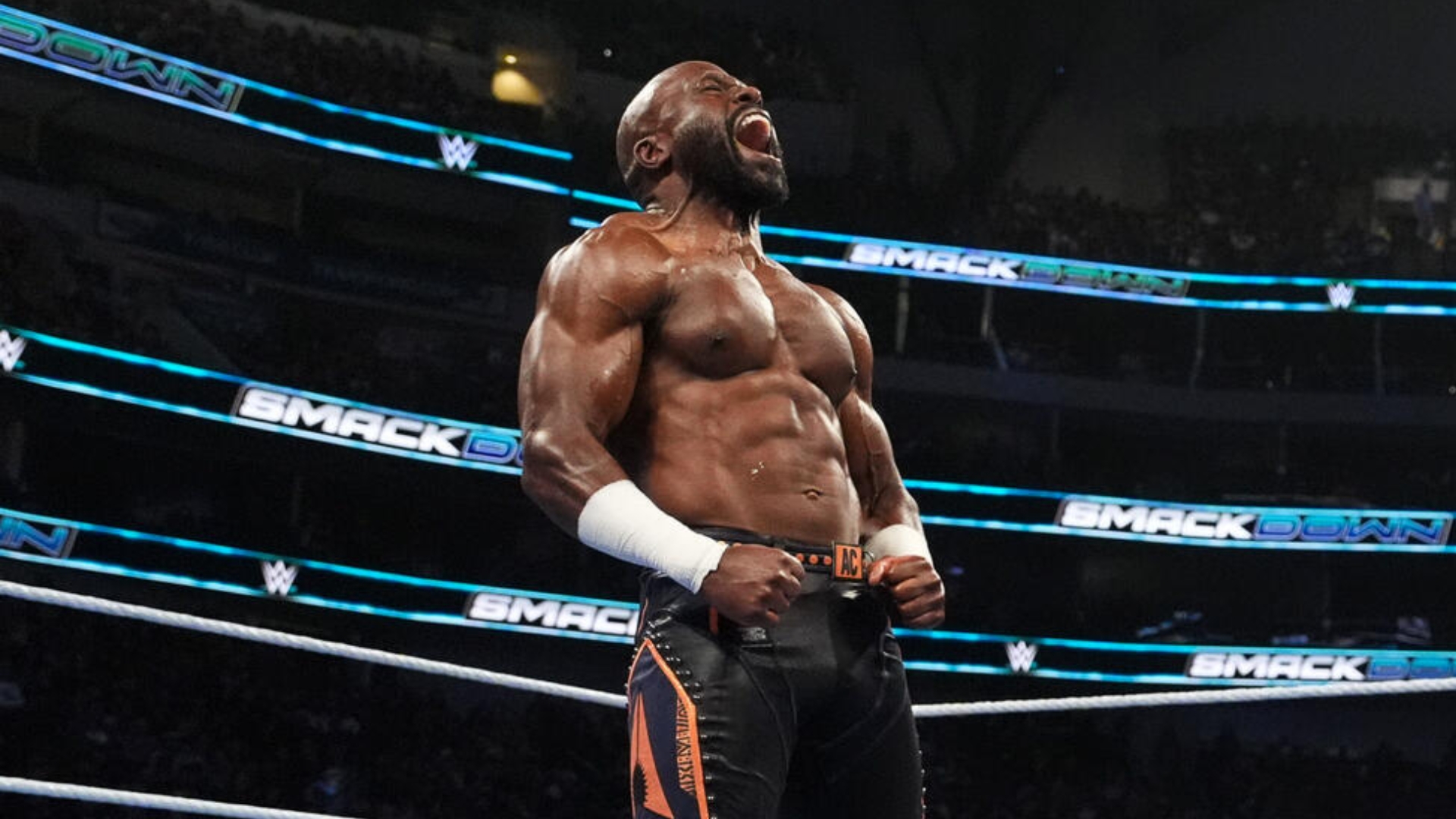 Muscular male wrestler in a WWE ring, fists clenched and shouting, wearing black/orange trunks and white wristbands, celebration pose.